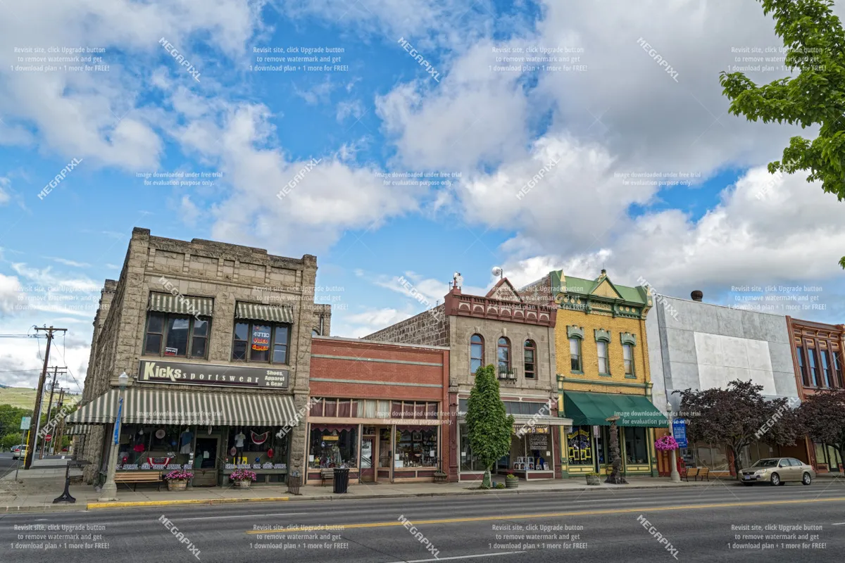 A lively downtown scene with colorful historic buildings, a Kicks sportswear storefront, tree-lined street, and flower planters. The area is bustling, with inviting storefronts and plenty of sidewalk activity, suggesting a vibrant local business district.