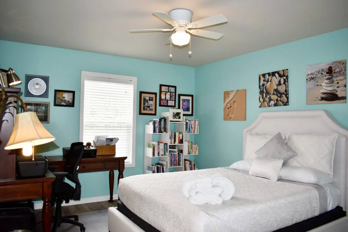 A bright bedroom that doubles as a dedicated workspace, featuring a bed, a large desk with chair, a bookshelf, and decorative artwork. The setting is conducive to both relaxation and productivity, with ample space and light for work or reading.
