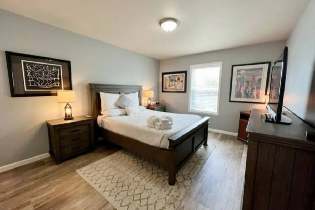 A serene bedroom with a queen-sized bed, a nightstand and lamp, softly colored bedding, artwork on the wall, and a window letting in gentle daylight. The overall decor is understated yet inviting, offering a restful place to recharge.