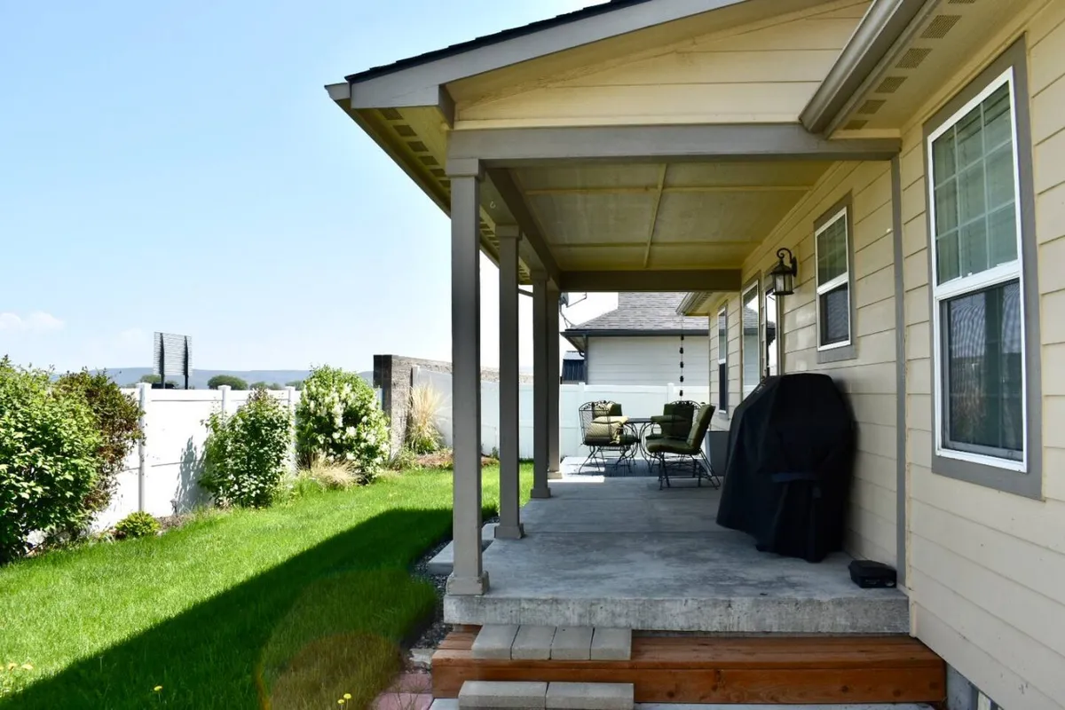This photo shows a covered porch with a green lawn, outdoor seating, and a grill, all set against a welcoming backdrop. The seating arrangement invites quiet mornings with coffee or lively evening barbecues, and the covered area provides shade and shelter.