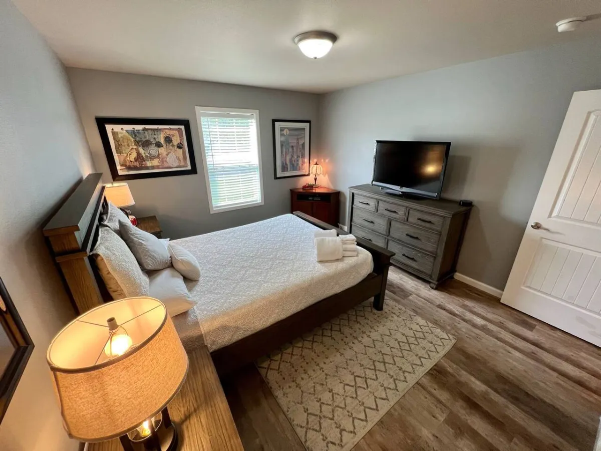 A cozy bedroom featuring a queen bed adorned with plush bedding, contemporary artwork on the walls, a nightstand with a lamp, and a flatscreen television positioned for comfortable viewing. Natural light filters in from a nearby window, creating a soothing atmosphere.