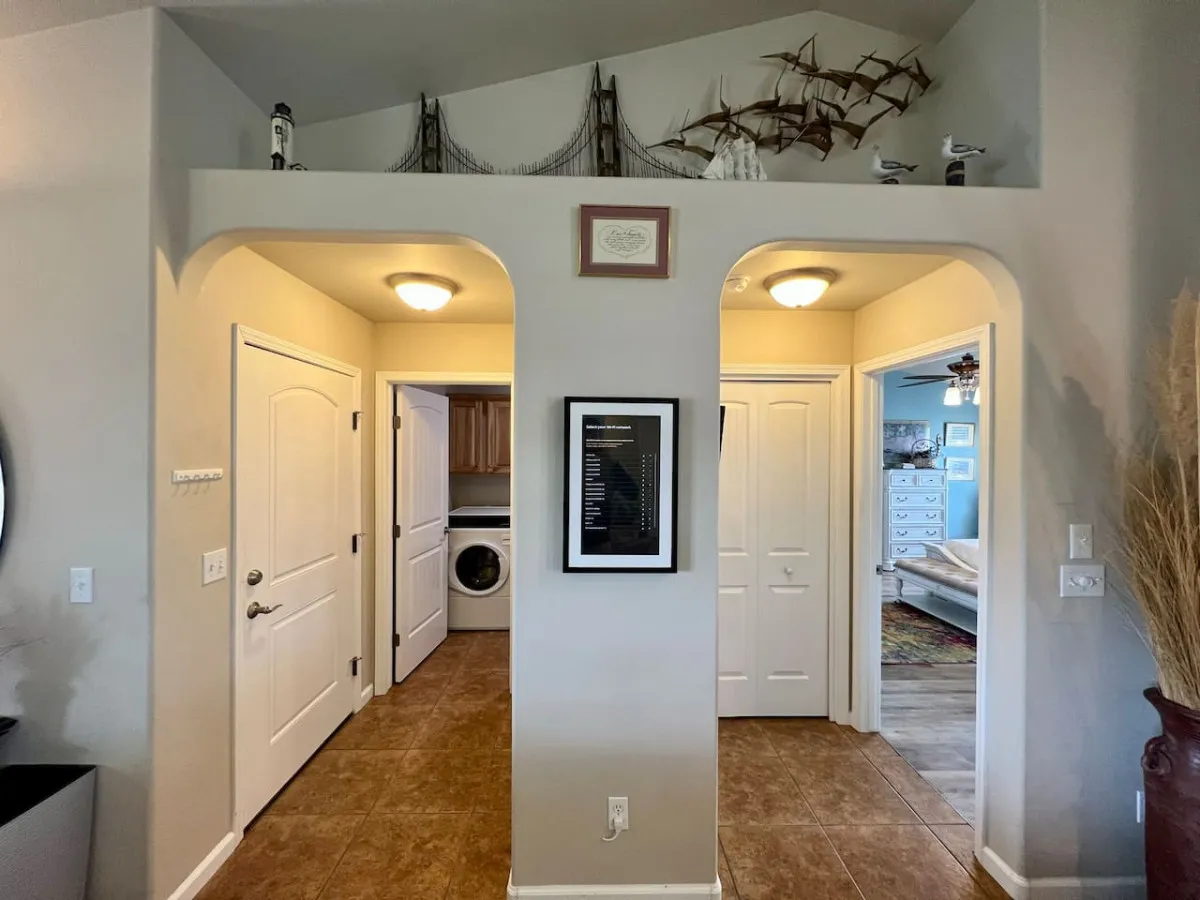 An inviting hallway scene with arched doorways, decorative ship models, a visible laundry area, and framed artwork. The perspective includes both entryway flow and a glimpse of the functional laundry space.