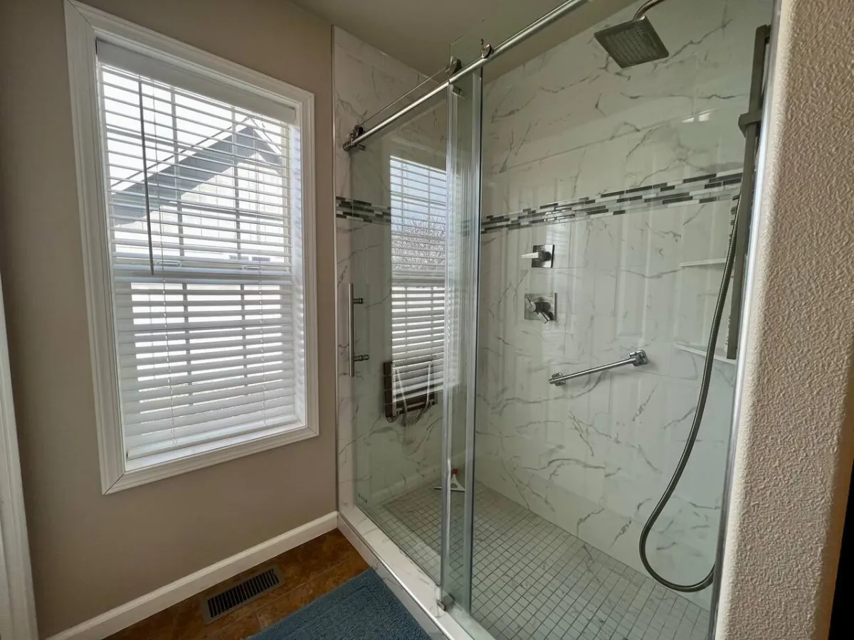 A modern, elegant bathroom with a glass shower enclosure, marble wall tiles, and sleek shower fixtures. Sunlight floods the space, highlighting the tranquility and luxurious finish ideal for self-care routines.